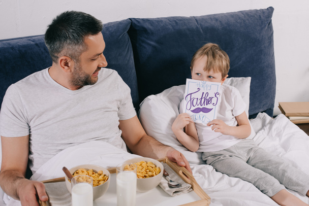 bonito menino cobrindo rosto com pais dia cartão de saudação enquanto sentado na cama perto do papai com café da manhã na bandeja
 - Foto, Imagem