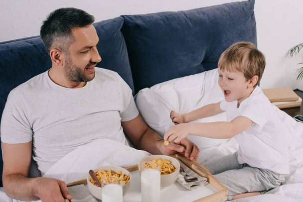 一緒にベッドに滞在しながら、陽気な父と息子が朝食を食べる  - 写真・画像