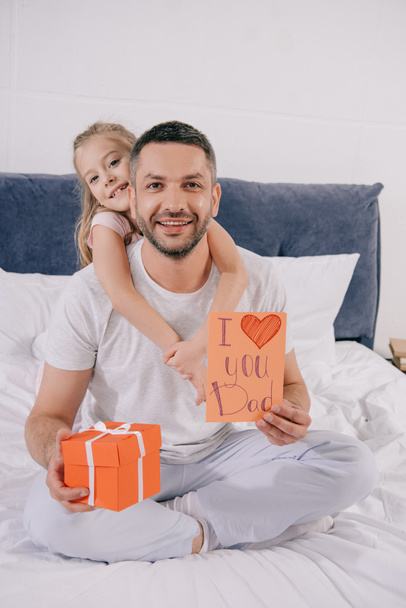 niño sonriente abrazando feliz papá sosteniendo caja de regalo y tarjeta de felicitación del día de los padres
 - Foto, Imagen
