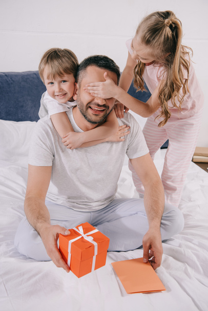 happy man holding gift box and fathers day greeting card while adorable kids hugging him and covering his eyes with hands - Foto, immagini