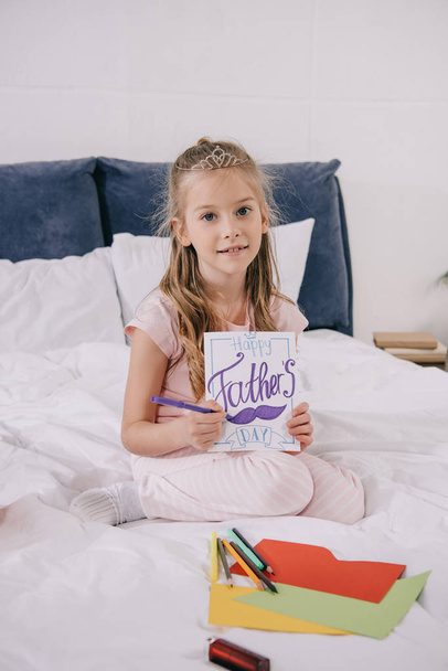 lindo niño dibujo padres día tarjeta de felicitación mientras está sentado en la cama y mirando a la cámara
 - Foto, imagen