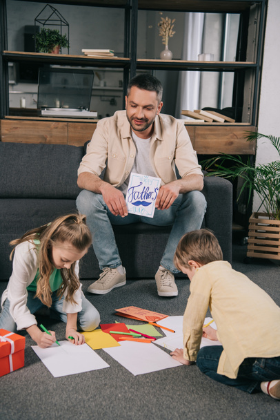 glücklicher Mann, der auf dem Sofa sitzt und eine Grußkarte zum Vatertag in der Nähe von Kindern hält, die auf Papierbögen zeichnen, während sie auf dem Boden sitzen - Foto, Bild