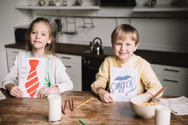 lachende broer en zus tonen vaders dag wenskaarten terwijl zittend aan keuken tafel met ontbijt geserveerd - Foto, afbeelding