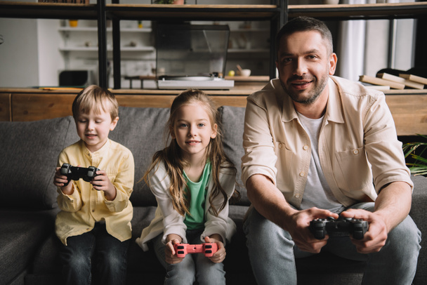 Kijów, Ukraina-10 maja 2019: szczęśliwy ojciec i urocze dzieci grając w gry wideo z joysticków w domu.  - Zdjęcie, obraz