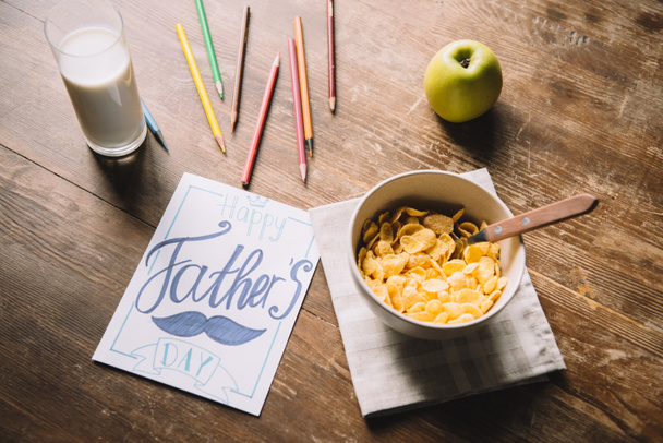 Открытки на день отца, разноцветные карандаши, миска с хлопьями, свежее яблоко и стакан молока на деревянном столе
 - Фото, изображение