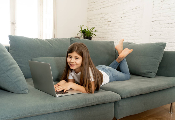 Beautiful cute charming schoolgirl playing and surfing the internet on laptop smiling and lying down on the couch at home. Digital technology, internet usage and life style concept. - Foto, Imagem