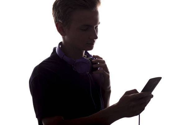 silhueta de menino ateenager com fones de ouvido em seu pescoço, um cara lançando através de uma lista de reprodução de música em seu dispositivo eletrônico em fundo isolado branco
 - Foto, Imagem