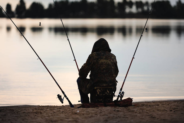 A pescar. Pescador pescando com três varas de pesca ao pôr do sol junto ao rio
  - Foto, Imagem