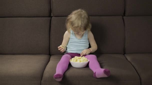 Girl sitting on sofa and eating corn puffs. Child smiling and taste puffcorns - Footage, Video