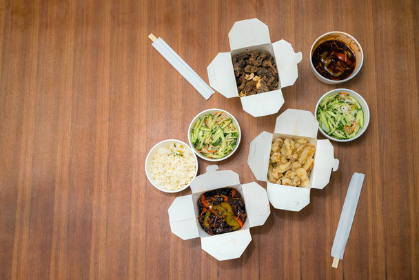 Top uitzicht van Chinees take-away eten met chop sticks op houten tafel. Pittige Aziatische gerechten in witte doos-salade, souse, rijst met ei, droog rundvlees, knapperige kip gekookt in olie. - Foto, afbeelding