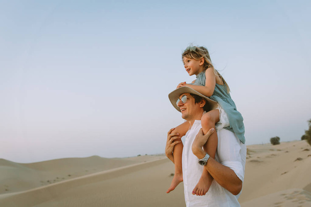 Niña sentada sobre los hombros de papá y mirando el atardecer en el desierto
 - Foto, Imagen
