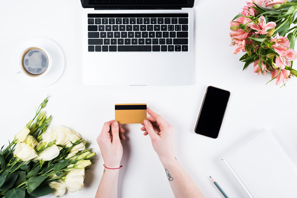 bijgesneden beeld van vrouw houden creditcard in de buurt van laptop, smartphone met blanco scherm, kopje koffie en boeketten op wit - Foto, afbeelding