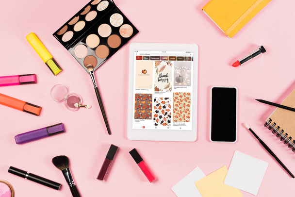 KYIV, UKRAINE - MAY 11, 2019: top view of digital tablet with pinterest app on screen, smartphone with blank screen, highlighters and decorative cosmetics on pink - Foto, Imagen