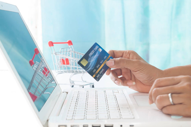 手はクレジットカードを保持し、ショッピングカートの背景を持つラップトップコンピュータを使用しています。オンラインショッピング、電子決済、インターネットバンキング - 写真・画像