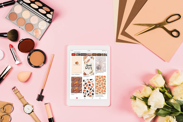 KYIV, UKRAINE - MAY 11, 2019: top view of digital tablet with pinterest app on screen, decorative cosmetics, flowers, craft paper and scissors on pink - Foto, Imagen
