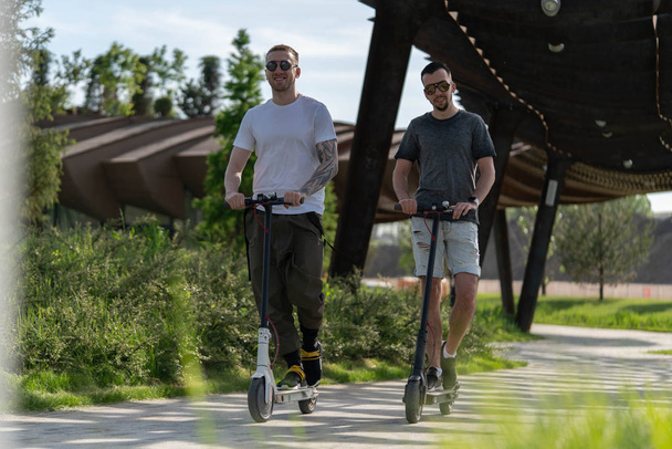 Two handsome men riding electric kick scooters at beautiful park landscape - Valokuva, kuva