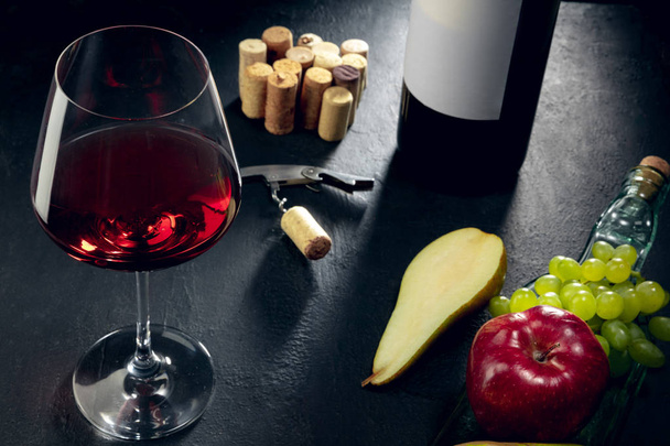 A bottle and a glass of red wine with fruits over dark stone background - Foto, Bild