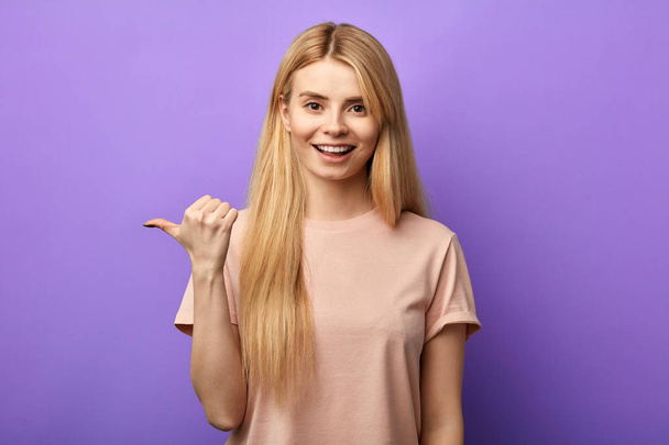 Optimistic young girl with toothy smile points aside with thumb - Фото, изображение