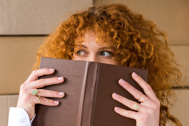 redhead girl covering face with book near cardboard boxes  - Photo, Image