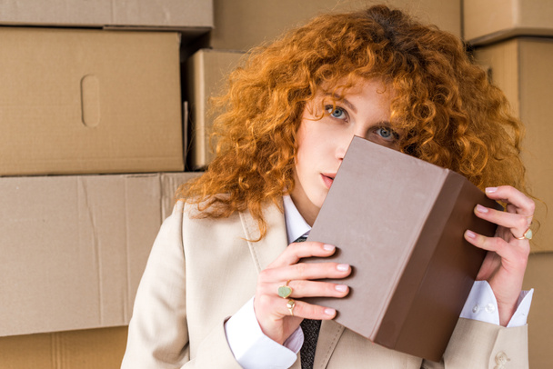 段ボール箱の近くに本で顔を覆う赤毛の女性  - 写真・画像