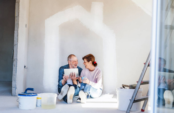 Senior couple painting walls in new home, using tablet. Relocation concept. - Zdjęcie, obraz