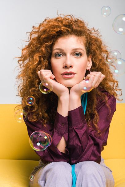 attractive curly redhead woman looking at camera and touching face near soap bubbles on white - Photo, Image