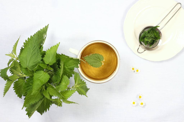 Healing tea with fresh nettles, flat view. Cup of nettle tea , fresh nettles, tea strainer on the plate,  light tablecloth   - Photo, Image