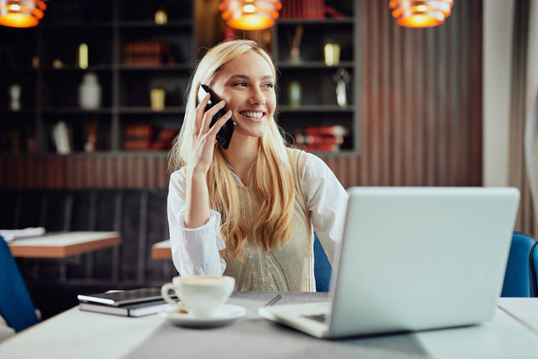 Νεαρή χαρούμενη Καυκάσια ξανθιά επιχειρηματίας ντυμένη έξυπνα casual κάθεται στο καφέ και χρησιμοποιώντας έξυπνο τηλέφωνο. - Φωτογραφία, εικόνα