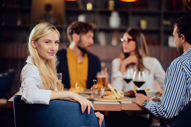 junge lächelnde kaukasische Blondine niedlich gekleidet elegant lässig am Tisch im Restaurant sitzend und über die Schulter schauend. Im Hintergrund ihre Freunde beim Abendessen. - Foto, Bild