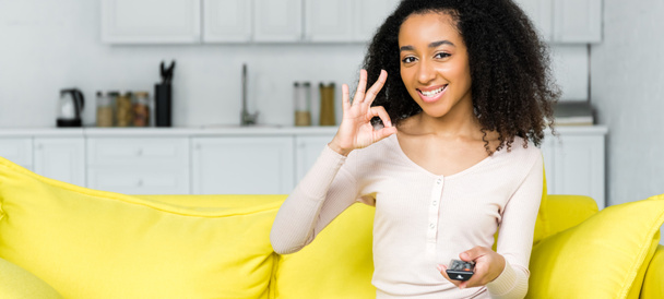 mujer afro-americana feliz con mando a distancia en la mano mostrando signo ok
 - Foto, Imagen