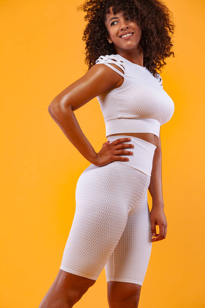 Smiling strong athletic woman with black skin and curly hair, doing exercise on white background wearing sportswear. Fitness and sport motivation. - Fotó, kép