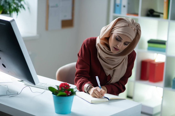 Beautiful Muslim woman writing into notebook while working - Foto, afbeelding