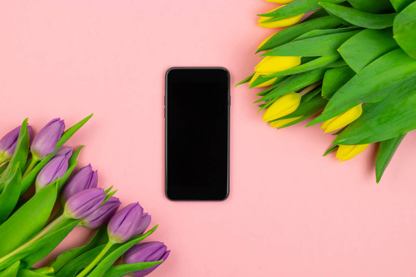 Bouquet of purple and yellow tulips and black smartphone on the pink background. Top view. Copy space. Presentation template. - Zdjęcie, obraz