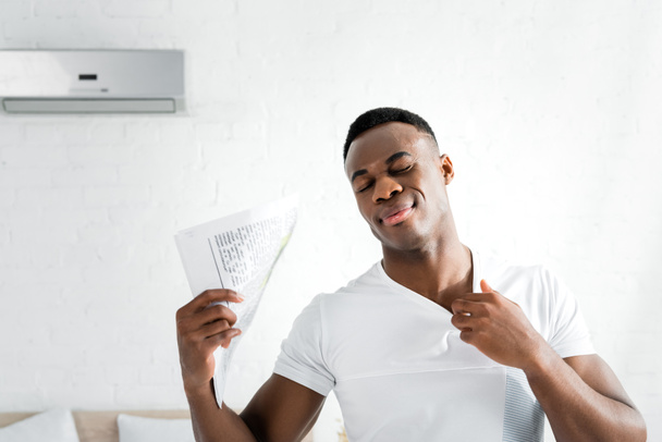 hombre afroamericano sudoroso cerrando los ojos, de pie en habitación blanca con temperatura de calor, sosteniendo periódico en la mano
 - Foto, Imagen