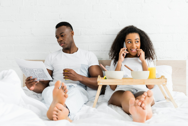 african american couple using smartphone, reading newspaper and lying in bed - Foto, Bild