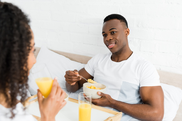 enfoque selectivo de hombre afroamericano con tazón de desayuno mirando a la novia, acostado en la cama
 - Foto, Imagen