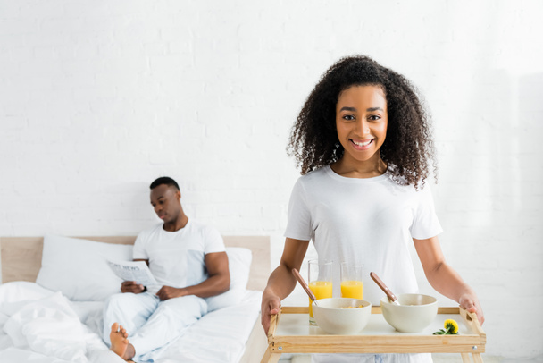 cheerful african american woman holding tray in hands, with boyfriend in bed on background - Fotoğraf, Görsel