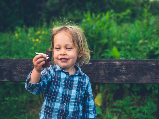 A little toddler is sitting on a bench in nature and is eating a fruit bar - Photo, image