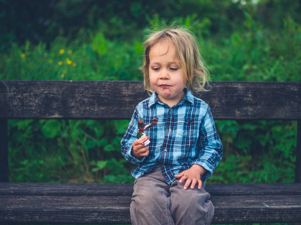 A little toddler is sitting on a bench in nature and is eating a fruit bar - Photo, Image