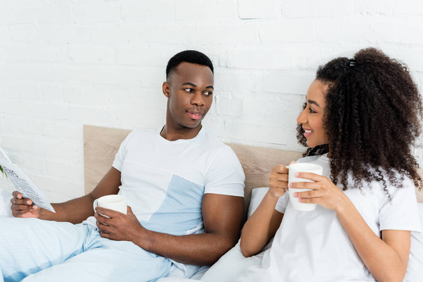 happy african american couple with newspaper and cup lying in bed, looking at each other - Photo, Image
