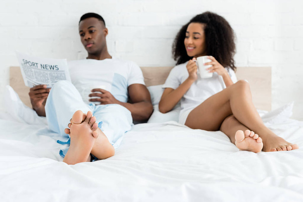 african american woman lying on bed near man with cup and newspaper - Photo, image