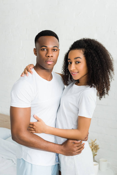 african american couple cuddling to each other, standing in white room, looking at camera - Photo, Image