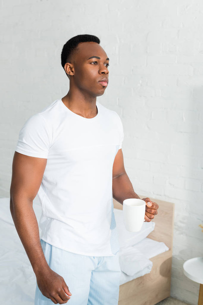 serious african american man standing in white room, holding cup with beverage in hand - Foto, Imagem