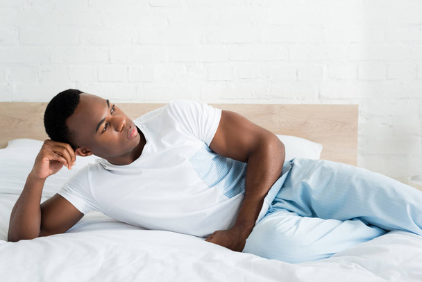 calm african american man looking away, laying on bed in white room - Foto, Imagem