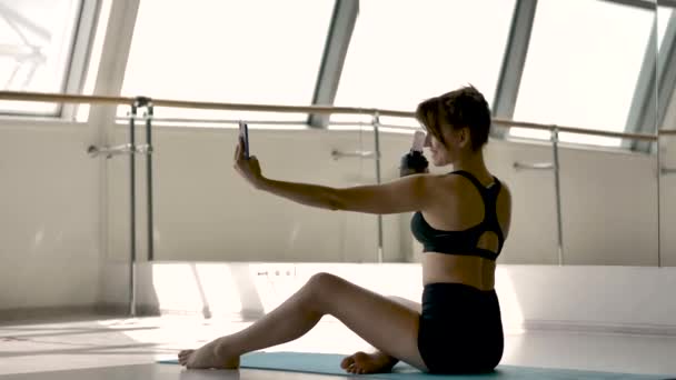 Caucasian sporty girl in sportswear is photographing herself on the phone while sitting on the floor in the gym. She is holding a shaker in her hand, there is a mirror in the background - Filmmaterial, Video