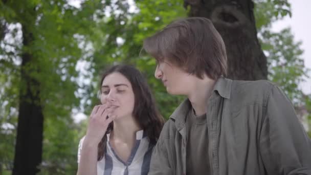 Portrait young happy couple in casual clothes spending time together in the park, having a date. Lovers sitting on the bench, leaning to each other. The boy looks in camera, winks, shows OK sign - Záběry, video