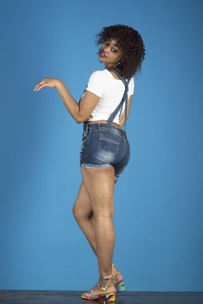 Lady with makeup looks back. Woman with african appearance in denim clothes looks gorgeous and stylish on blue background. Fashion concept. Lady in modern clothes as short denim overalls - Foto, Imagen