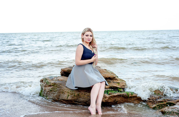 Blonde woman in stylish dress sitting on rocks by sea - Фото, изображение