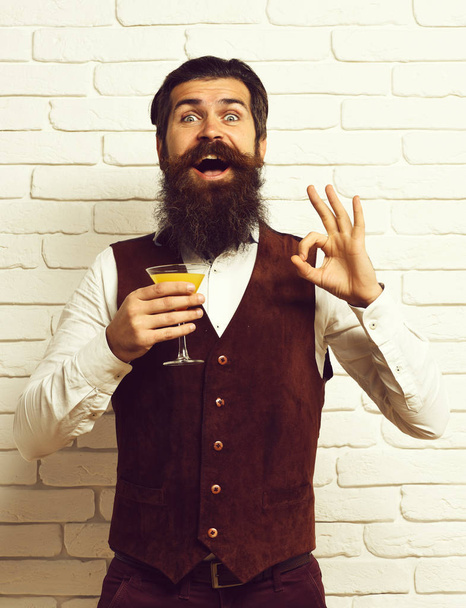 handsome bearded man with long beard and mustache has stylish hair on smiling face holding glass of alcoholic beverage in vintage suede leather waistcoat on white brick wall studio backgroun - Valokuva, kuva