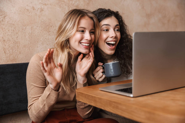 Feliz chicas guapas amigos sentados en la cafetería usando ordenador portátil saludando hablando
. - Foto, Imagen